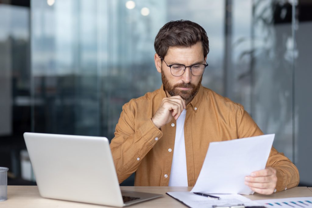 A man in glasses reviews documents while working on his laptop in an office setting.